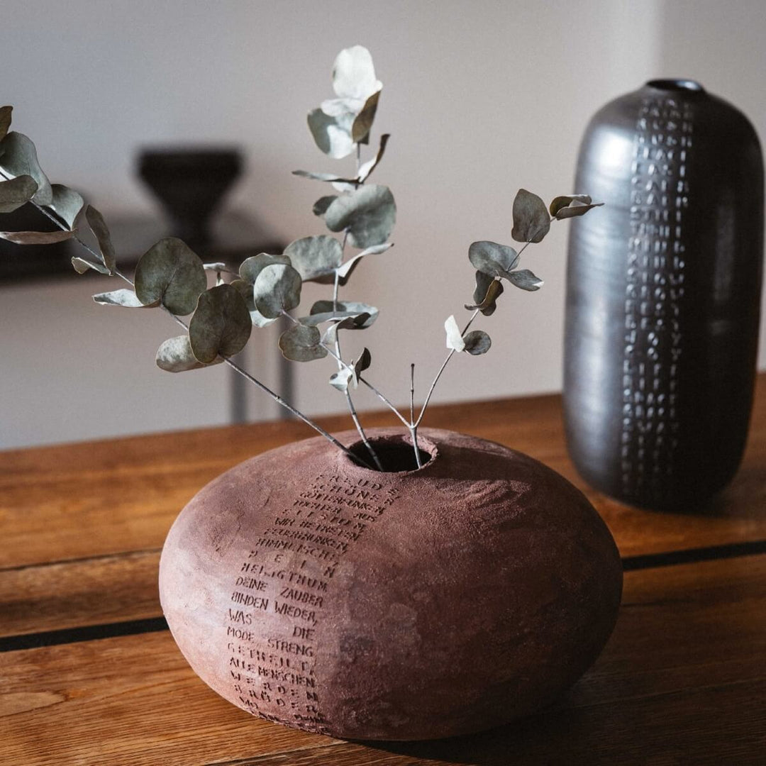 Vase aus Keramik, rund und bauchig aus rotem Steinzeug Ton mit Schillers Ode "And die Freude" als Relief von oben nach unten verlaufend auf der Vase angebracht. In der Vase steckt ein Eukalyptus Zweig. | haptik